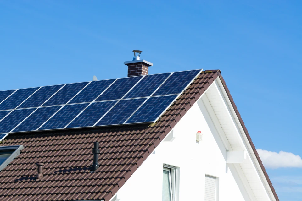 Gros plan sur un toit de maison avec des panneaux solaires au dessus, sur un ciel bleu avec des nuages et des rayons du soleil